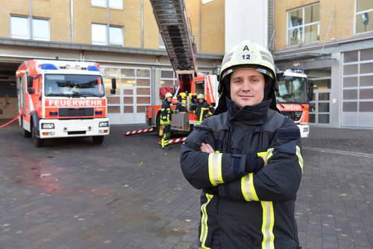 Portrait of confident firefighter in front of fire engine with colleagues in background