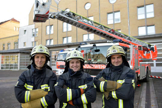 Portrait of three smiling firefighters standing on yard in front of fire engine