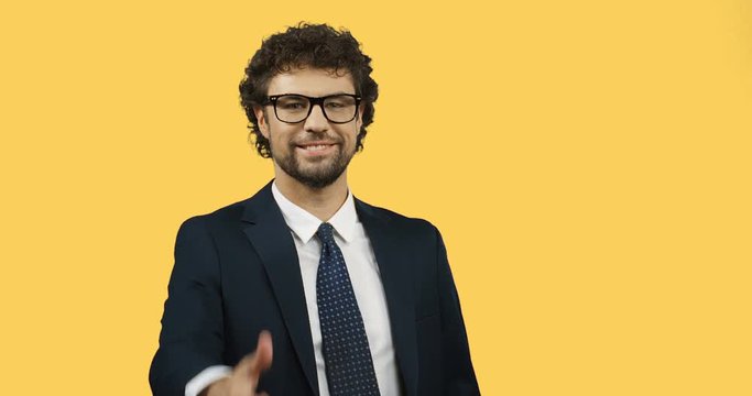 Handsome cheerful businessman in glasses, suit and tie giving his thumb up while standing on the yellow screen background.