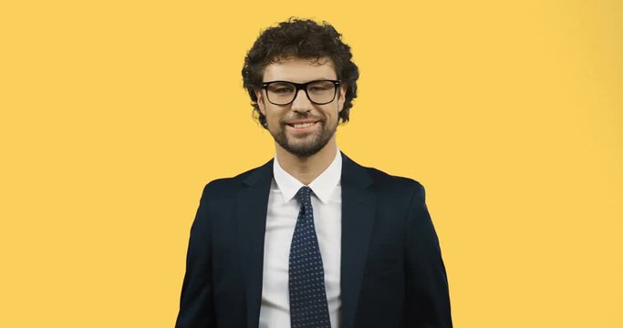 Good looking smiled man in glasses, suit and tie doing ok gesture with fingers while standing on the yellow screen background.