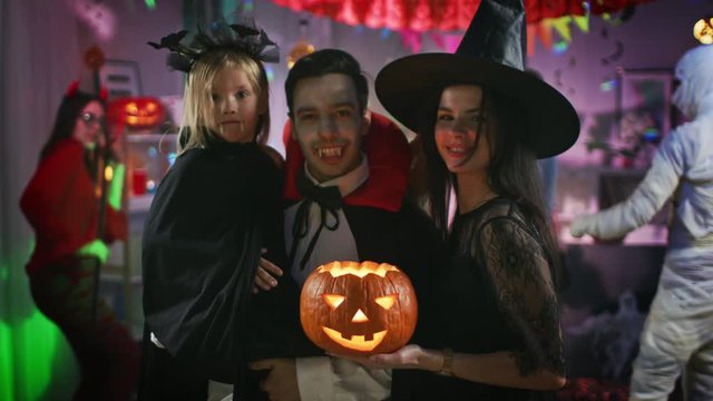 Halloween Costume Party: Father Count Dracula Holds Little Bat Girl Daughter and Hugs Witch Wife for a Happy Family Portrait. In the Background Monsters Having Fun