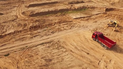 View of the empty large red truck moving on a sandy track near the yellow excavator in desert or dunes. Beautiful view from above.