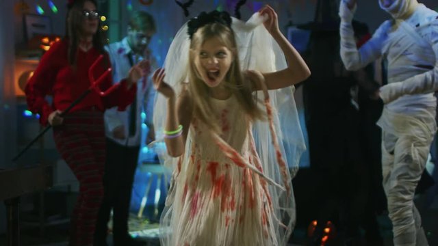 Halloween Costume Party: Little Girl in a Bloody White Bride Dress Turns Around and Makes Scary Faces. In the Background Group of Monsters Dancing and Having Fun in Decorated Room with Disco Lights