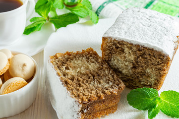 Slices of biscuit cake, a сup of tea, small cookies and mint leaves on a white wooden table.