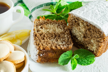 Slices of biscuit cake, a сup of tea with lemon, small cookies and mint leaves on a white wooden table.