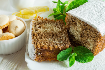Slices of biscuit cake, slice of lemon, small cookies and mint leaves on a white wooden table.