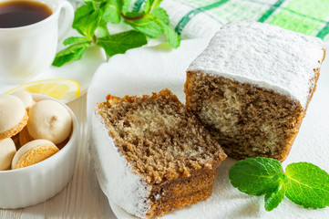 Slices of biscuit cake, a сup of tea with lemon, small cookies and mint leaves on a white wooden table.