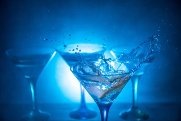Martini cocktail glass in hand splashing on dark toned smoky background or colorful cocktail in glass with splashes and olives. Party club entertainment. Mixed light. Selective focus