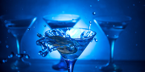 Martini cocktail glass in hand splashing on dark toned smoky background or colorful cocktail in glass with splashes and olives. Party club entertainment. Mixed light. Selective focus