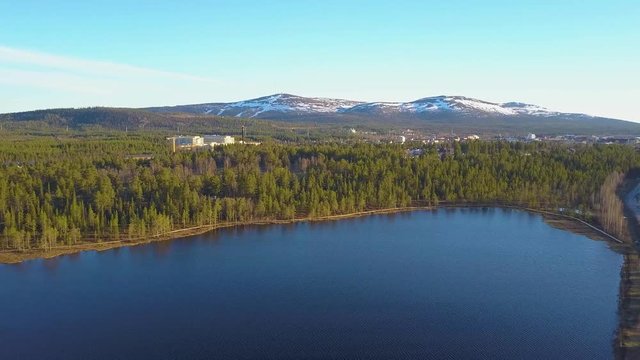 Aerial, tracking, drone shot, of a tornetrask lake, forest, a town and snowy, fell mountains, in the background, Swedish wilderness, on a sunny, spring day, in Abisko, Kiruna, Lapland, North Sweden