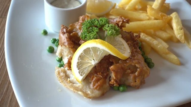Close up panning shot across fish, chips & peas dinner, supper on white plate, wooden table with slice of lemon garnish.