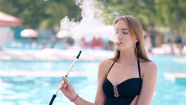 CU, Slow motion, Portrait: Young beautiful girl is sitting on the beach in summer, on a deckchair, and smokes a hookah.