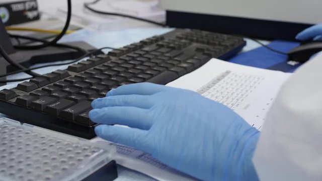Doctor or a scientist is working on his research on the computer, cross checking he data on a paper sheet.