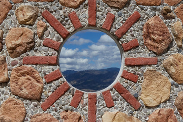 wall ornamented with stone and bricks simulating a sun, with a hole through which mountains and clouds can be seen