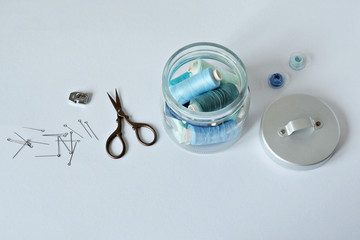 Blue thread, glass jar, bobbins, scissors, zipper foot and metal pins over white