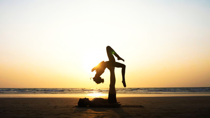 Silhouette of fit sporty couple practicing acrobatic yoga with partner together on the beach in...