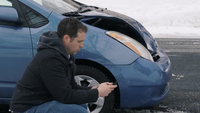 Man Squats Next To Wrecked Car And Takes Pictures. camera remains fixed as crouched man takes pictures of the front passenger side of his wrecked car.