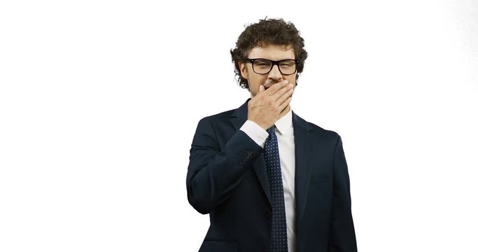 Portrait of the attractive Caucasian businessman in glasses, suit and tie yawning while standing on the white wall background.