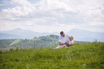 Fototapeta na wymiar Happy father with his little son on his knees sits against a background of green forest, mountains and sky with clouds. Friendship concept