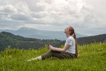 Fototapeta na wymiar Beautiful dreaming girl with a laptop sitting on green grass on a background of mountains