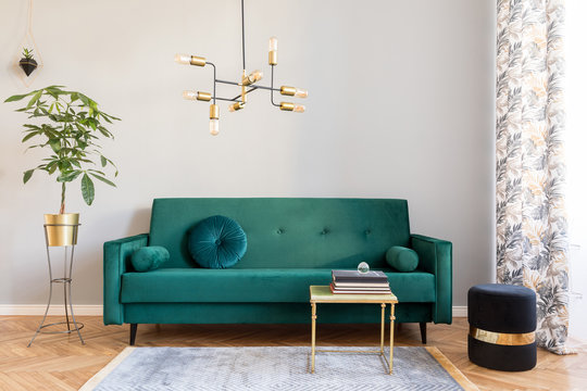 Bright and sunny minimalistic home interior with design green velvet sofa, furniture,plant, gold coffee table with books and elegant pendant lamp. Brown wooden parquet. Stylish decor of living room.