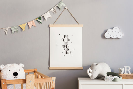 Stylish and cute scandinavian decor of newborn baby room with mock up poster, natural toys, hanging decor flags and cloud, wooden cradle, plush rhino  and teddy bears. Grey walls.