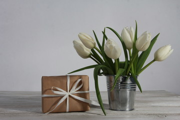Bunch of white tulips in tin bucket and gift box on white wooden table. Greeting concept. White ribbon, women or mothers day, spring, side view