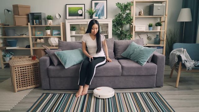 Beautiful Asian girl is turning on robotic vacuum cleaner then sitting on sofa and using smartphone enjoying convenience of modern technology. People and devices concept.