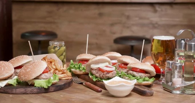 Juicy meat inside fresh loafs of breed with vegetables and sauce on side. A big luch prepared for a group of people. Burgers on wooden surface with beer and pickles.