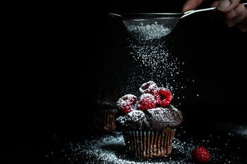 dark, romantic photo of a chocolate muffin, decorated with fresh, red raspberries. Above her a woman's hand is holding a small silver sieve and sprinkles with powdered sugar