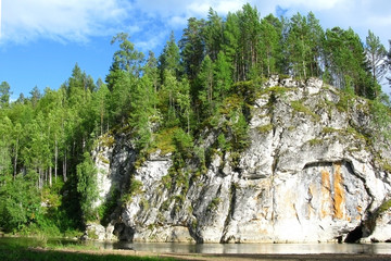 Steep cliff above the river with forest on top. Bright Sunny day, calm water flow.