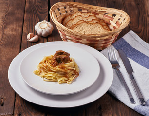Tagliatelle with thick veal sauce in a white plate, next to garlic, bread, cutlery and napkin on a dark wooden background, close-up