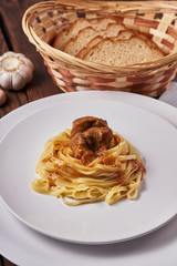 Tagliatelle with thick veal sauce in a white plate, next to garlic, bread on a dark wooden background, close-up
