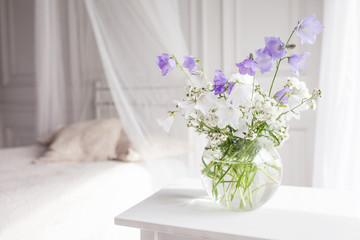 Glass vase with lilac and white floweers  in light cozy bedroom interior. White wall, bed with white linen, light blanket or plaid and pillows.