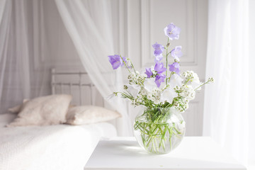 Glass vase with lilac and white floweers  in light cozy bedroom interior. White wall, bed with white linen, light blanket or plaid and pillows.