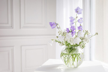 Glass vase with lilac and white floweers  in light cozy bedroom interior. White wall, sunlight from window, copy space