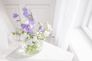 Glass vase with lilac and white floweers  in light cozy bedroom interior. White wall, sunlight from window, copy space