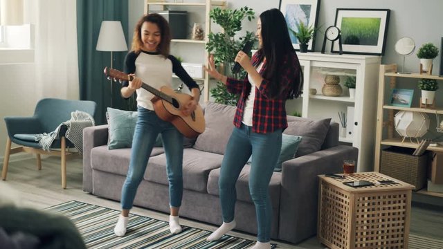 Cute young ladies Asian and African American are singing in remote control and playing the guitar relaxing at home expressing positive emotions. Friendship and fun concept.