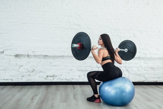 Close-up photo of confident young woman doing weight lifting workout at gym turning back. Attractive young woman bodybuilder lifting barbells looking focused and sitting on blue ball.