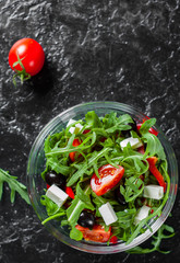 fresh green salad with arugula, tomatoes, cheese, pepper and olive in glass bowl on dark background