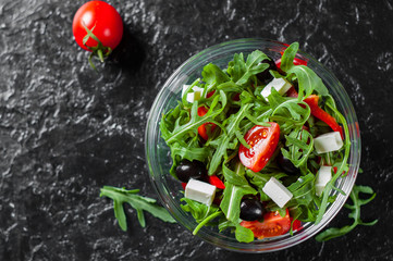 Fototapeta na wymiar fresh green salad with arugula, tomatoes, cheese, pepper and olive in glass bowl on dark background