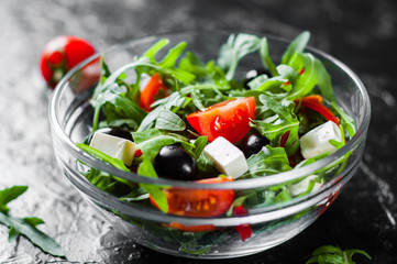 fresh green salad with arugula, tomatoes, cheese, pepper and olive in glass bowl on dark background