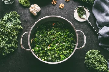 Poster Stewed kale in cooking pot on dark kitchen table background with ingredients and spoon, top view © VICUSCHKA