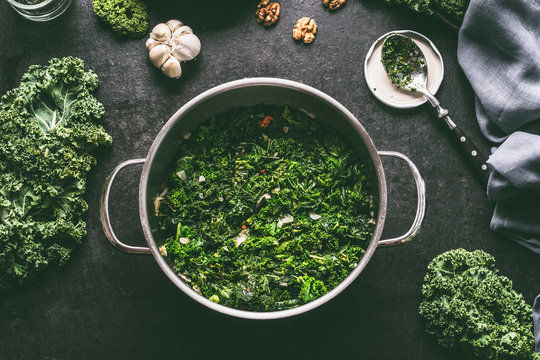 Stewed kale in cooking pot on dark kitchen table background with ingredients and spoon, top view