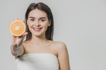A young beautiful girl stands on a gray background. During this, it smiles genuinely and holds one hand in a horizontal position while holding half an orange.