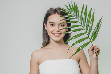 Fototapeta premium A beautiful young girl in the studio on a white background with wet skin and wet hair holds a large green tropical leaf in hands and covers a part of her face.
