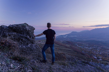 Fototapeta premium A young man in a black T-shirt stands high in the mountains and looks at a purple sunset.