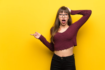 Woman with glasses over yellow wall surprised and pointing finger to the side