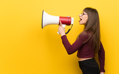 Woman with glasses over yellow wall shouting through a megaphone
