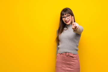 Woman with glasses over yellow wall showing and lifting a finger